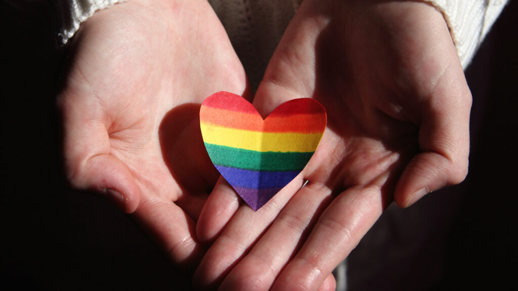 Caucasian hands holding a heart that has been colored with a rainbow.