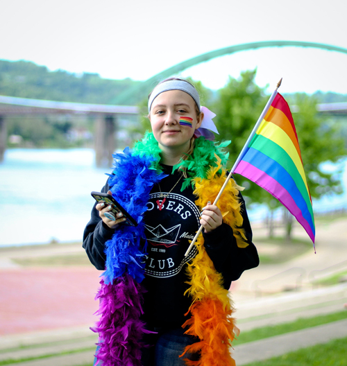 A person with a LGBTQIA+ pride flag. The person is also wearing a rainbow colored boa and black shirt and is holding a phone. They are smiling.