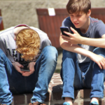 Two teen boys sit on a curb in an outdoor location. They are both looking at cell phones.