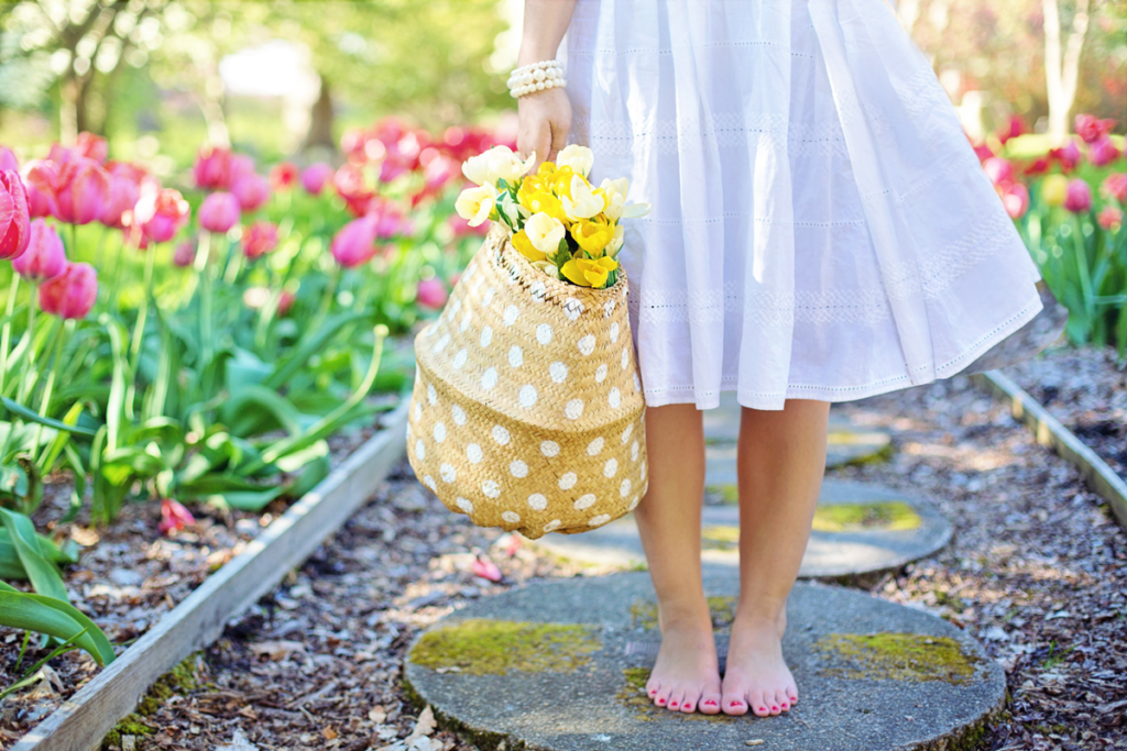 A woman in a white dress stands barefoot on a garden path. She is visible from the waste down only and holds a colorful basket of flowers in her right hand.