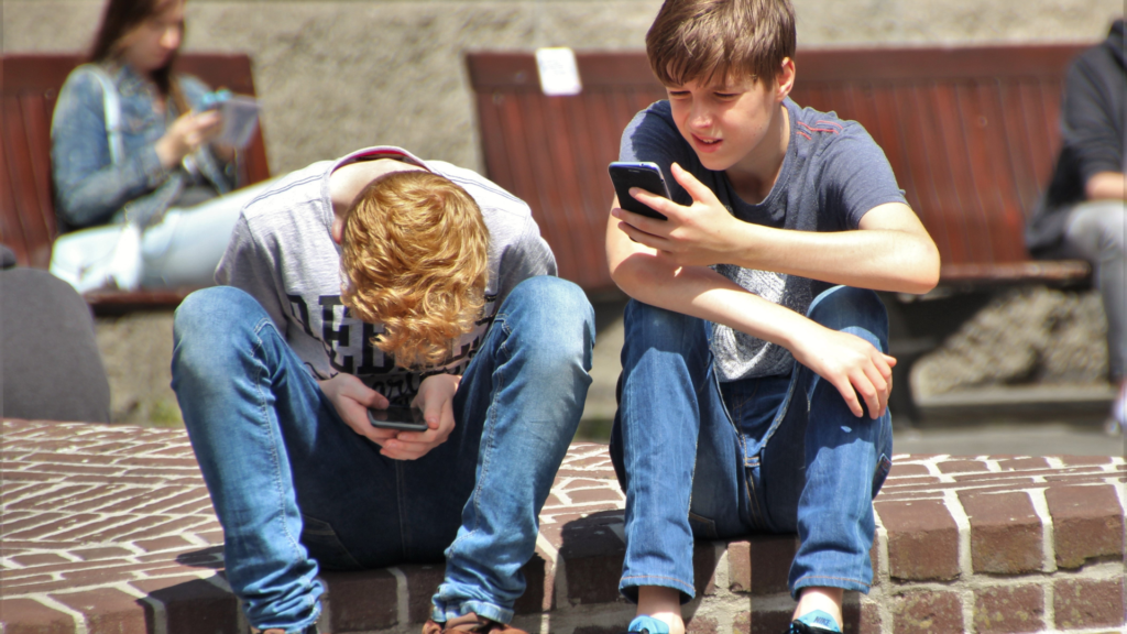 Two teen boys sit on a curb in an outdoor location. They are both looking at cell phones.